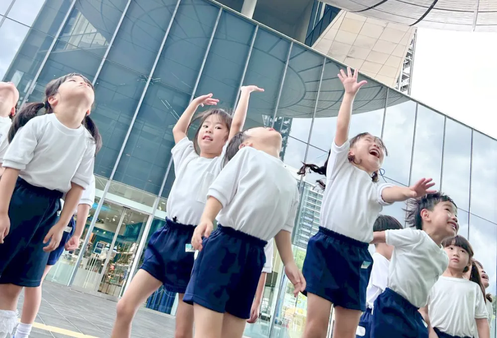 遠足で来た建物の前で空を見上げ、手を伸ばして喜ぶ子供たち