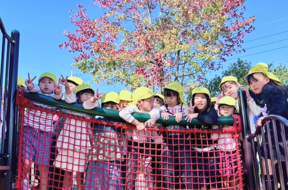 秋の木々の下、大型遊具のネットから笑顔を見せる園児たち