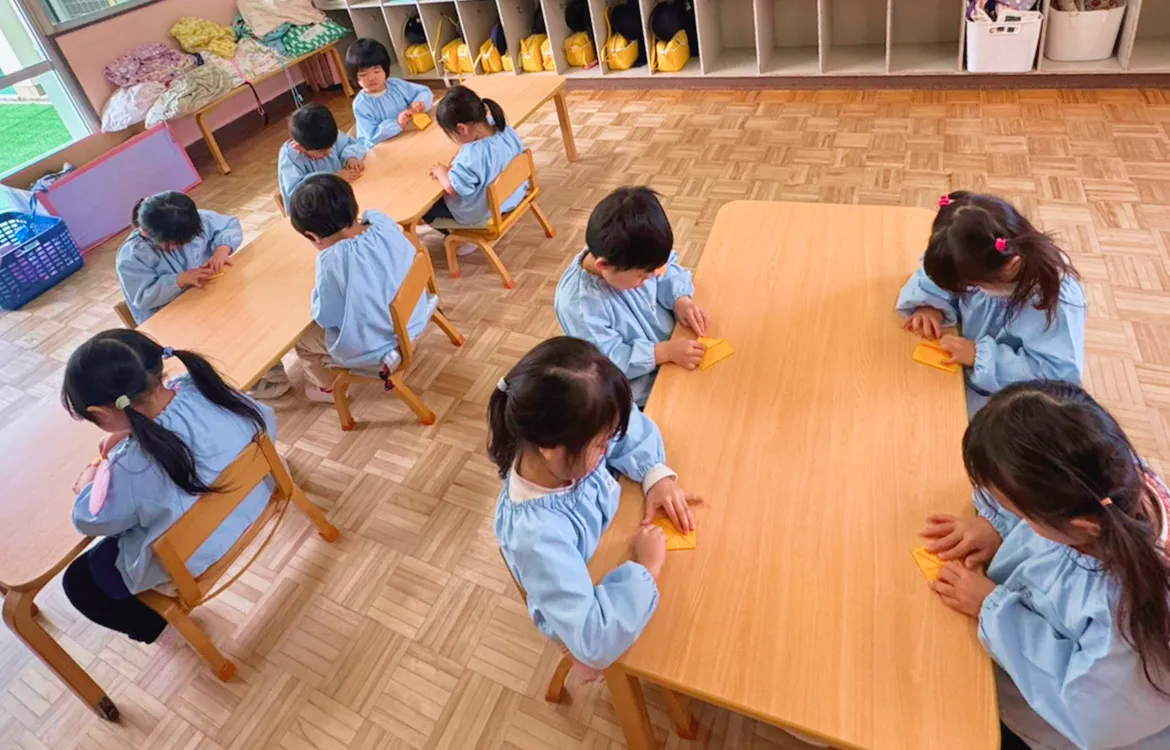 教室の机でお友達と一緒に集中して折り紙をする子供たち